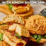 Close-up of gochujang chicken burgers with kimchi bacon jam on buns, garnished with greens and skewered, on a wooden board. Text overlay promotes a recipe from girlcarnivore.com.