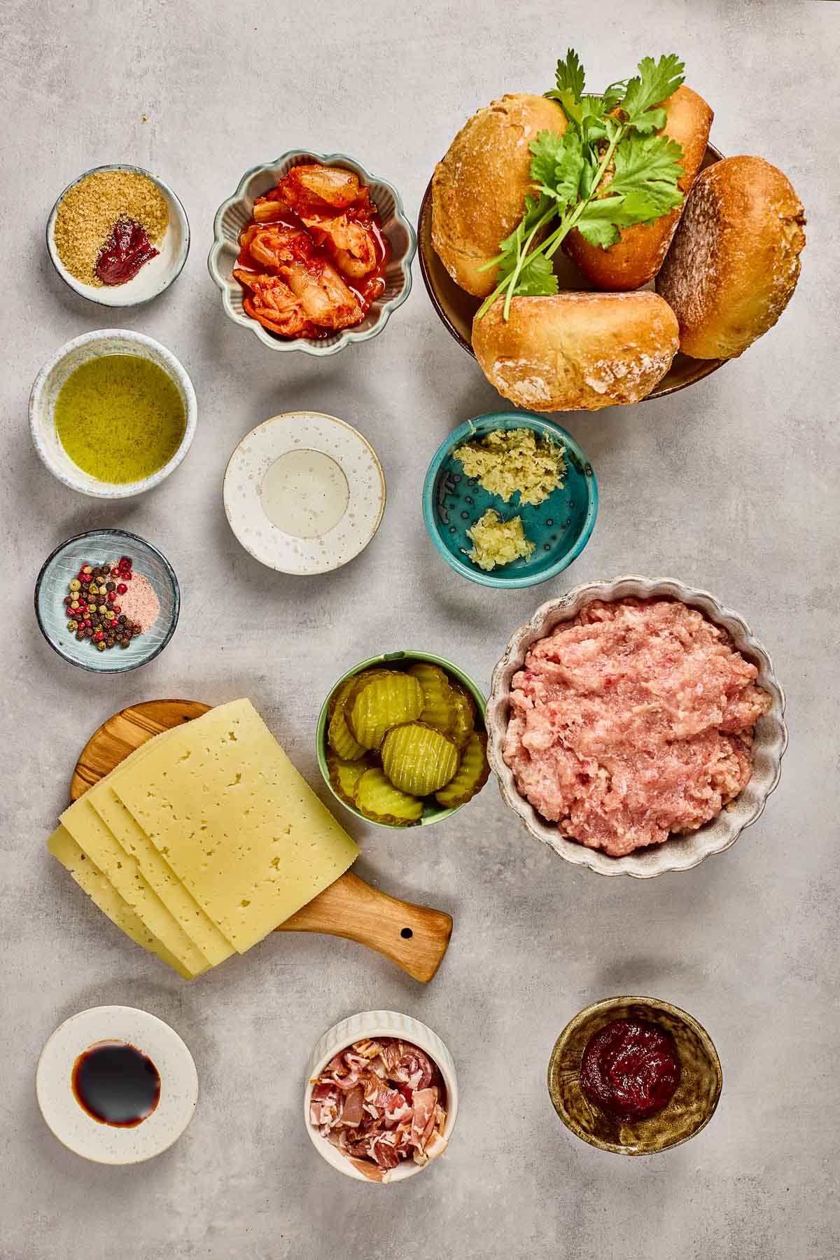 An overhead view of burger ingredients, including ground meat, bread rolls, cheese slices, pickles, sauces, seasonings, herbs, and small bowls of other condiments on a light surface.