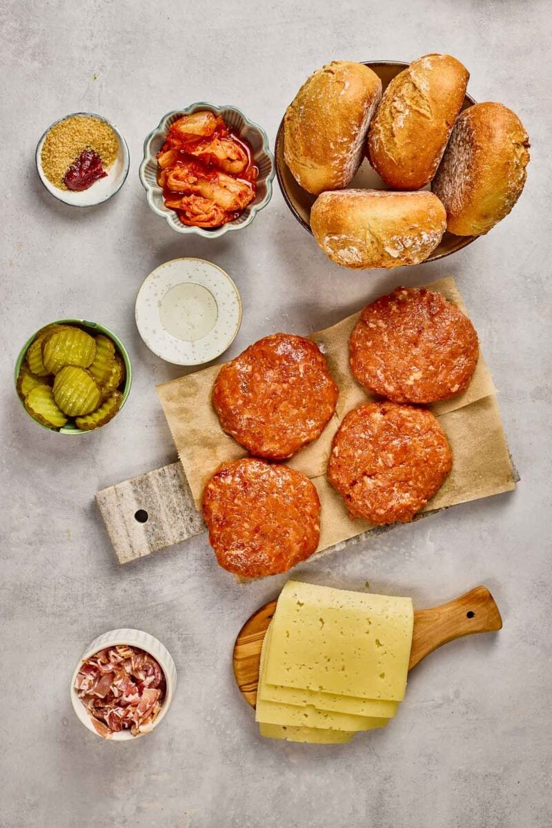 Top-down view of chicken burger ingredients including four raw patties, cheese slices, pickles, kimchi, buns, bacon, sauce, and oil arranged on a light surface.