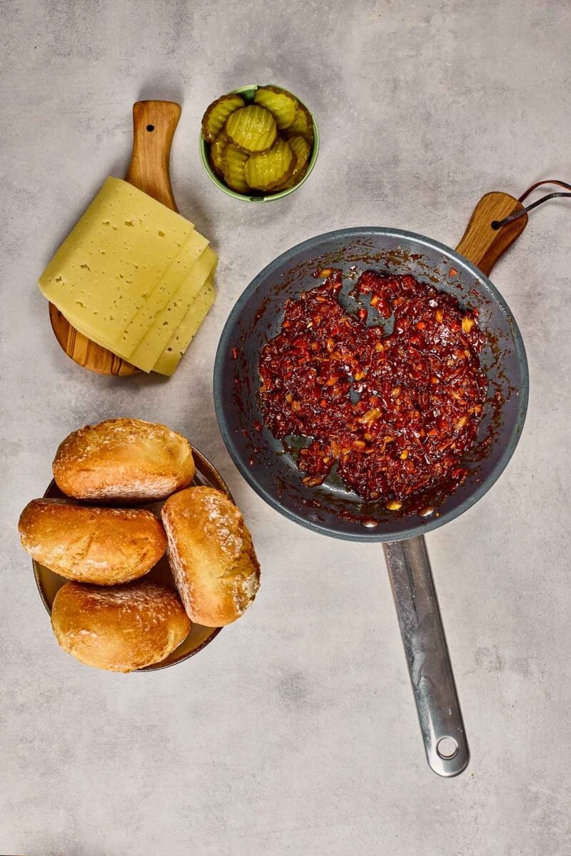 A pan with cooked bacon jam mixture, a plate of sandwich rolls, a stack of cheese slices on a wooden board, and a bowl of pickle slices on a gray background.