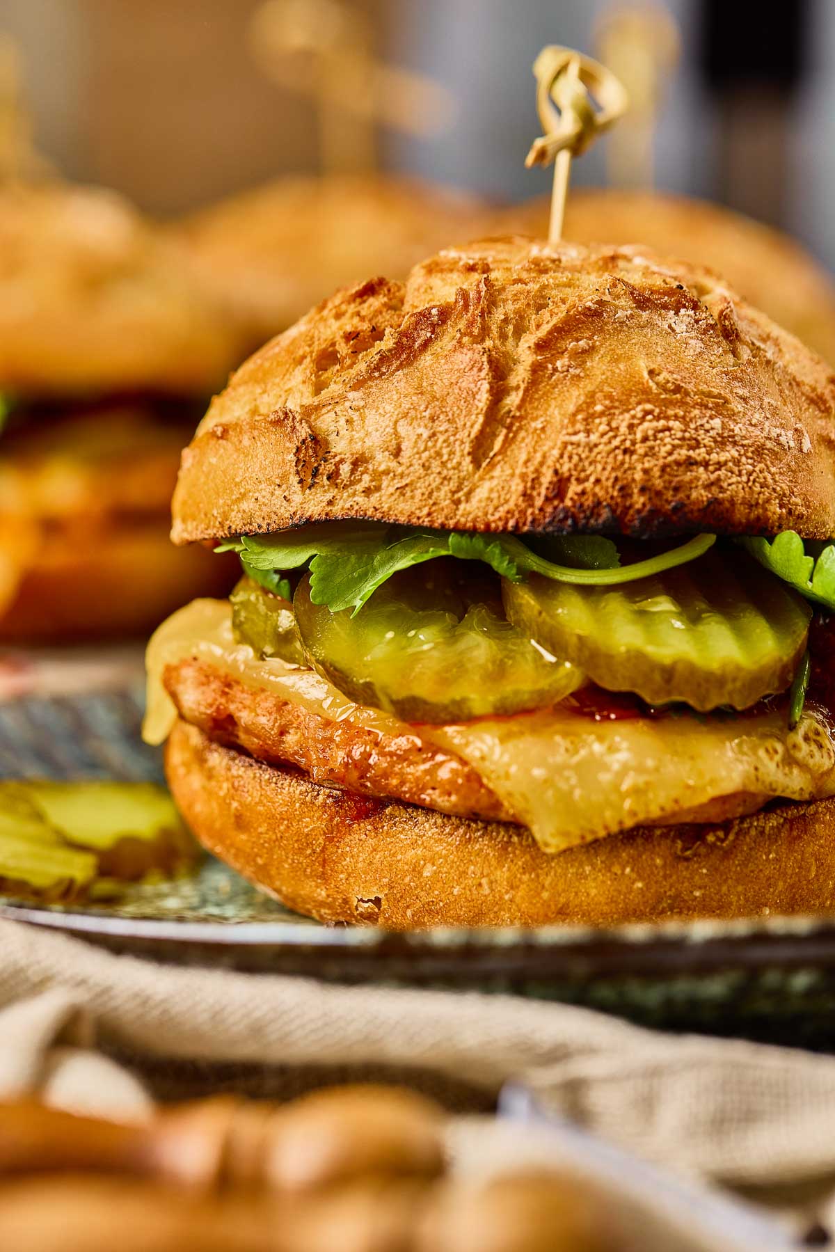 A close-up of a Gochujang Chicken Burger with a crusty bun, cheese, pickles, lettuce, and a cooked patty, secured with a toothpick. Another sandwich is blurred in the background.