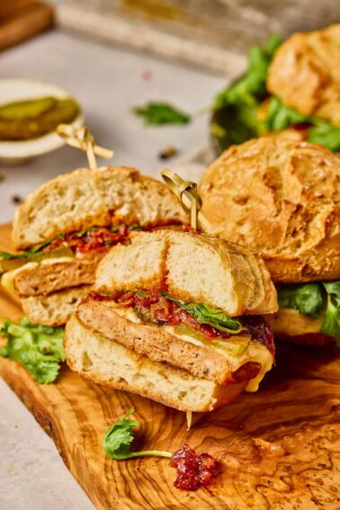 A Gochujang Chicken Burger with a chicken patty, cheese, greens, and kimchi bacon jam, cut in half and served on a wooden board with cilantro garnish.