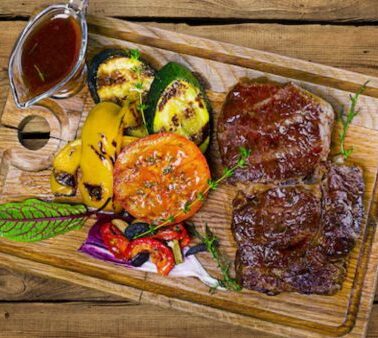 Grilled steak served on a wooden board with grilled vegetables, including tomato, zucchini, bell pepper, and a small dish of sauce.