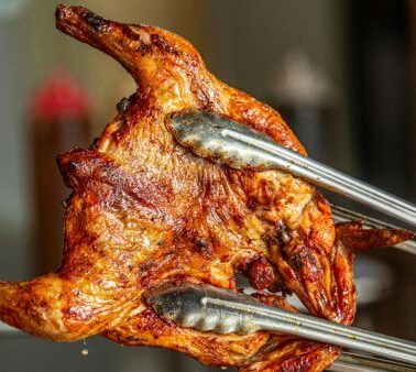 A person holds a whole roasted chicken with metal tongs, showing its crispy, golden-brown skin.