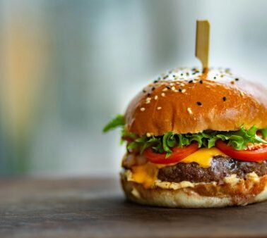 A cheeseburger with lettuce, tomato, cheese, and sauce in a sesame seed bun, held together with a wooden stick, sits on a wooden surface.