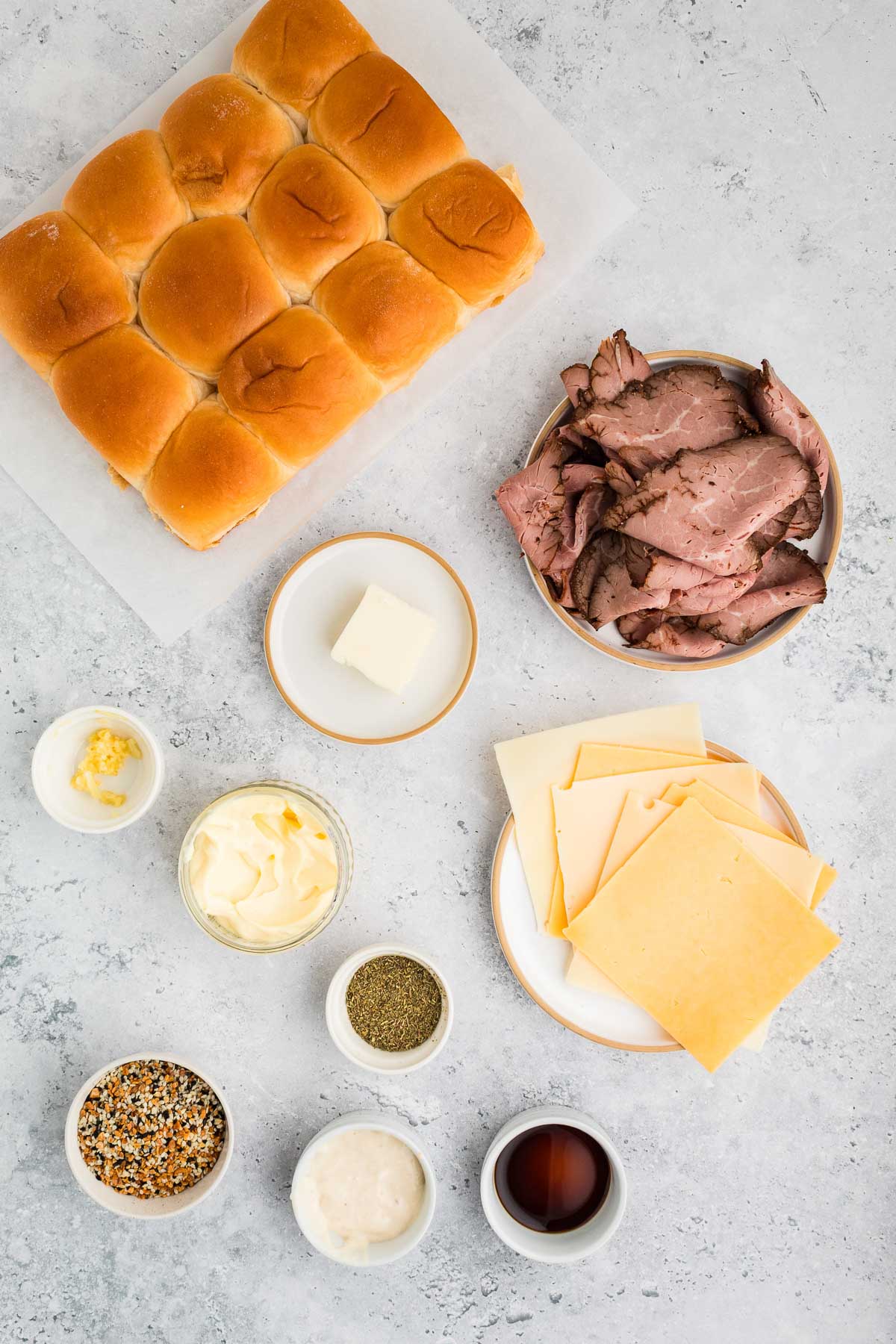 Ingredients for roast beef sliders arranged on a light surface, including slider buns, roast beef slices, cheese, butter, seasonings, mayonnaise, and sauces in small bowls.
