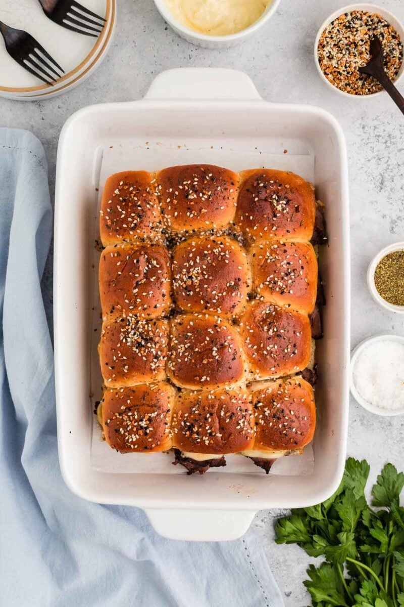A white baking dish contains twelve golden-brown slider buns topped with sesame seeds, surrounded by bowls of spices, plates, and a blue napkin.