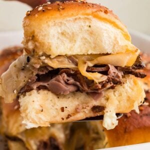 A hand holding a roast beef slider with melted cheese, and a soft bun, with more sandwiches in a dish below.