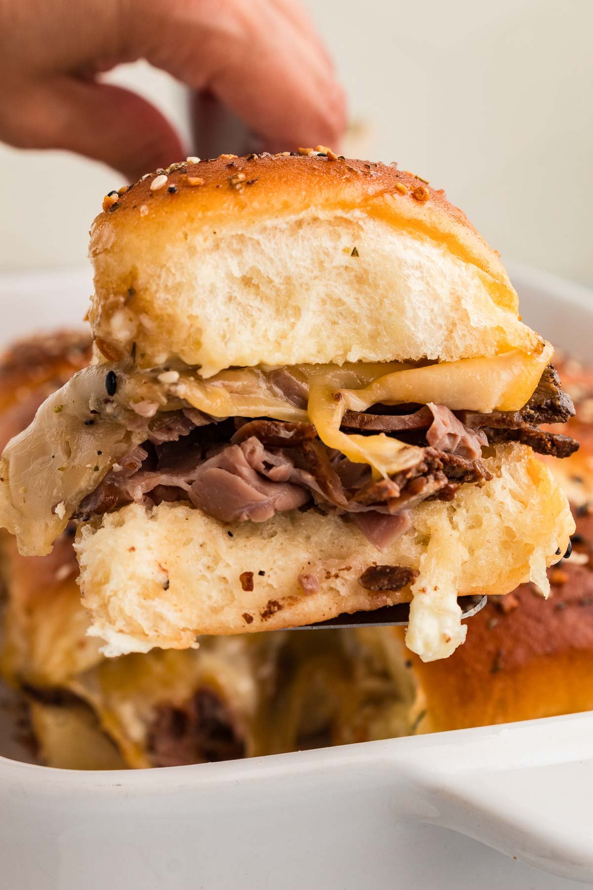 A hand holding a roast beef slider with melted cheese, and a soft bun, with more sandwiches in a dish below.