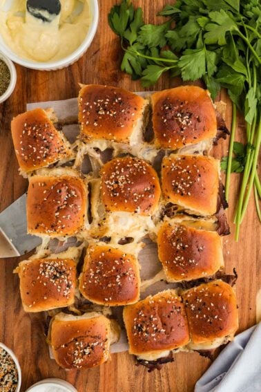 A batch of 12 golden-brown roast beef sliders with sesame topping sits on parchment paper, surrounded by parsley and condiments on a wooden surface.