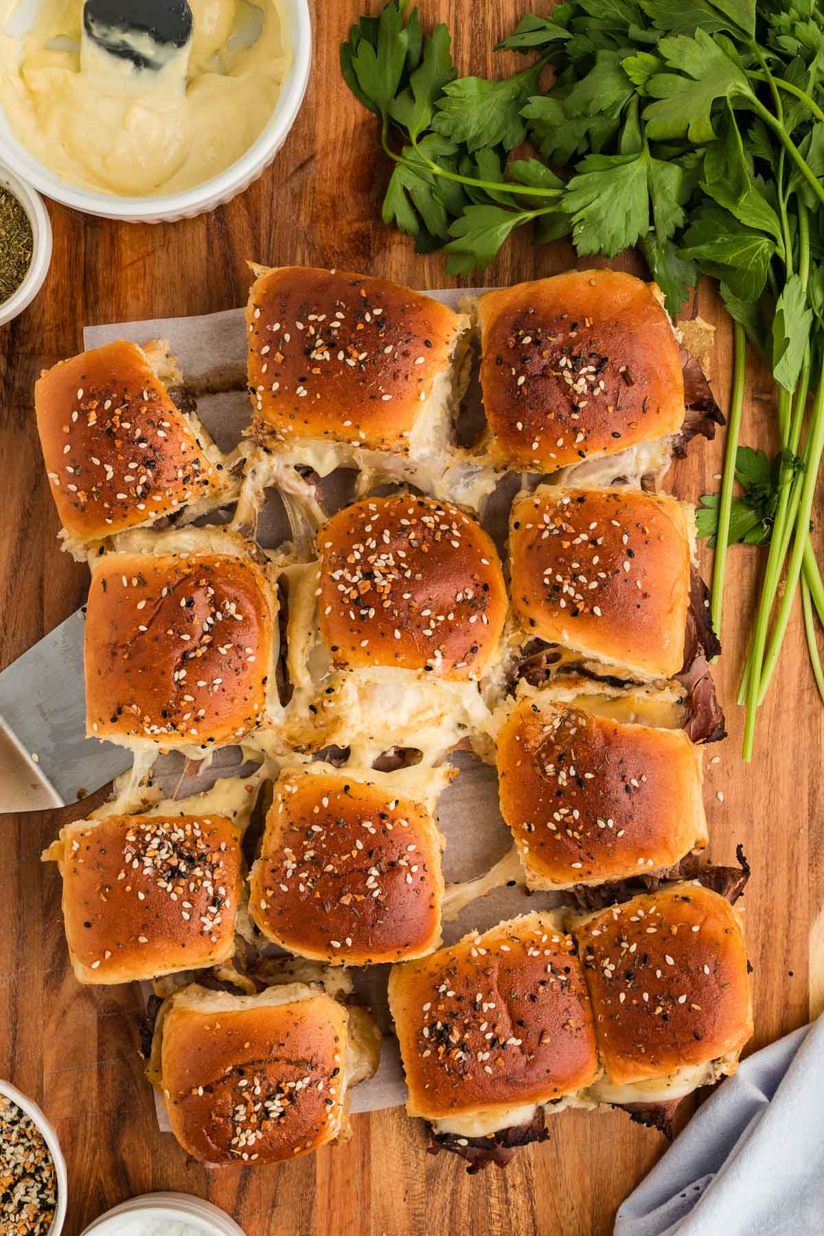 A batch of 12 golden-brown roast beef sliders with sesame topping sits on parchment paper, surrounded by parsley and condiments on a wooden surface.