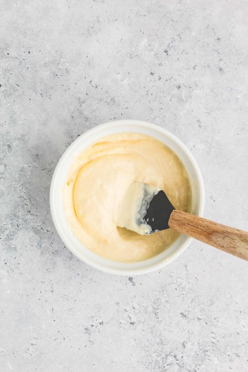 A small white bowl filled with creamy horseradish sauce sits on a light gray surface, with a black spatula featuring a wooden handle resting inside.