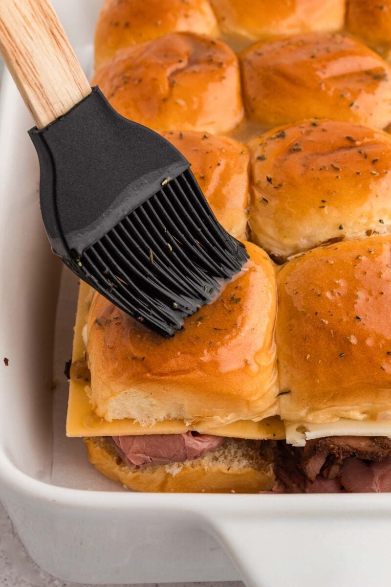 A silicone brush is spreading butter on top of slider buns in a white baking dish.