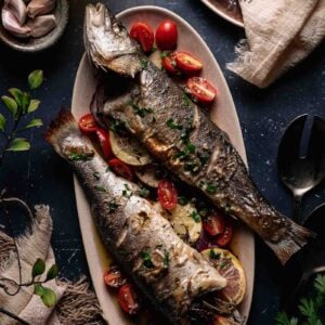 Two whole roasted branzino are served on a platter with lemon slices, halved cherry tomatoes, and chopped herbs, surrounded by tableware and ingredients on a dark surface.