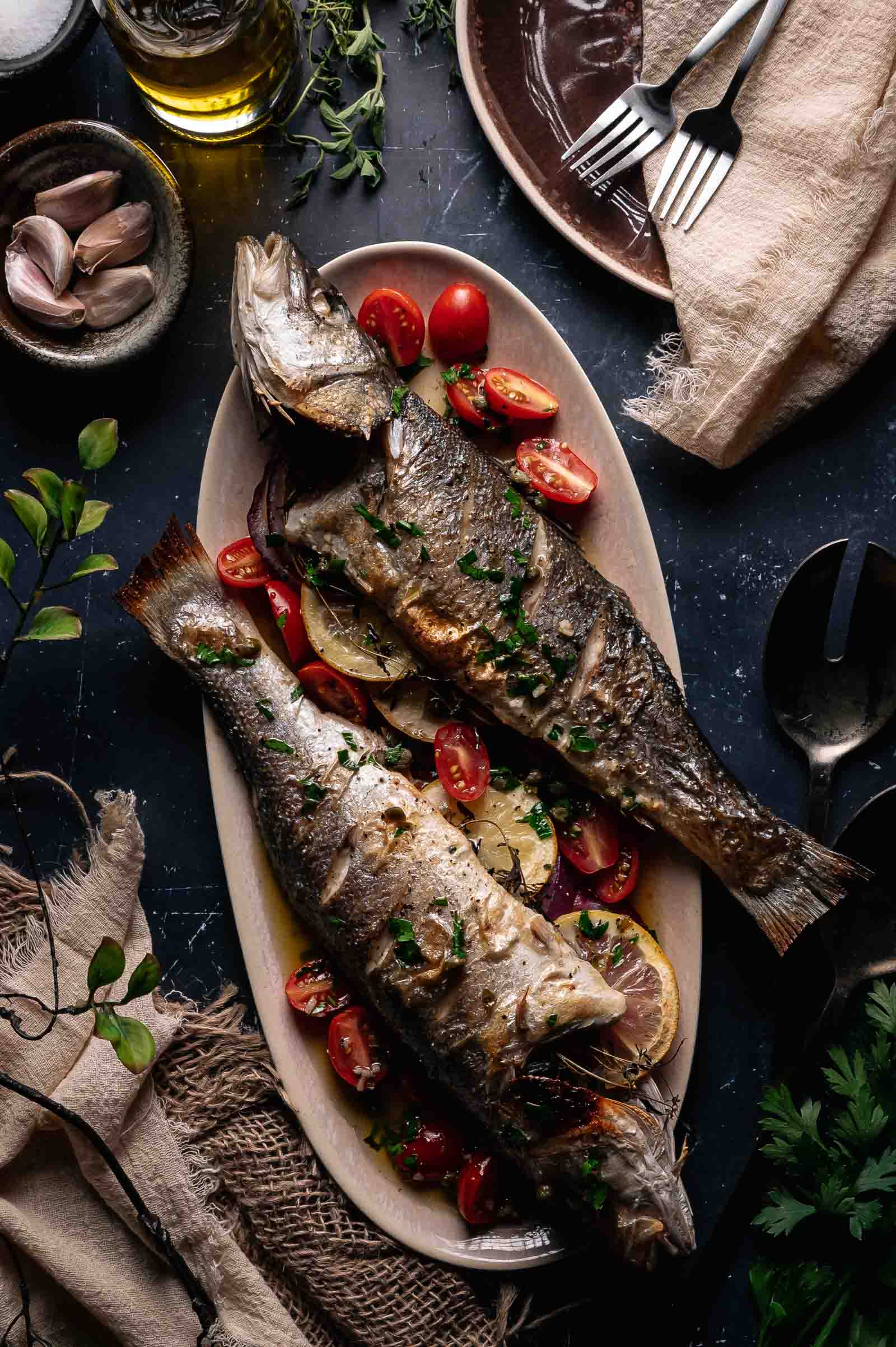 Two whole roasted branzino are served on a platter with lemon slices, halved cherry tomatoes, and chopped herbs, surrounded by tableware and ingredients on a dark surface.