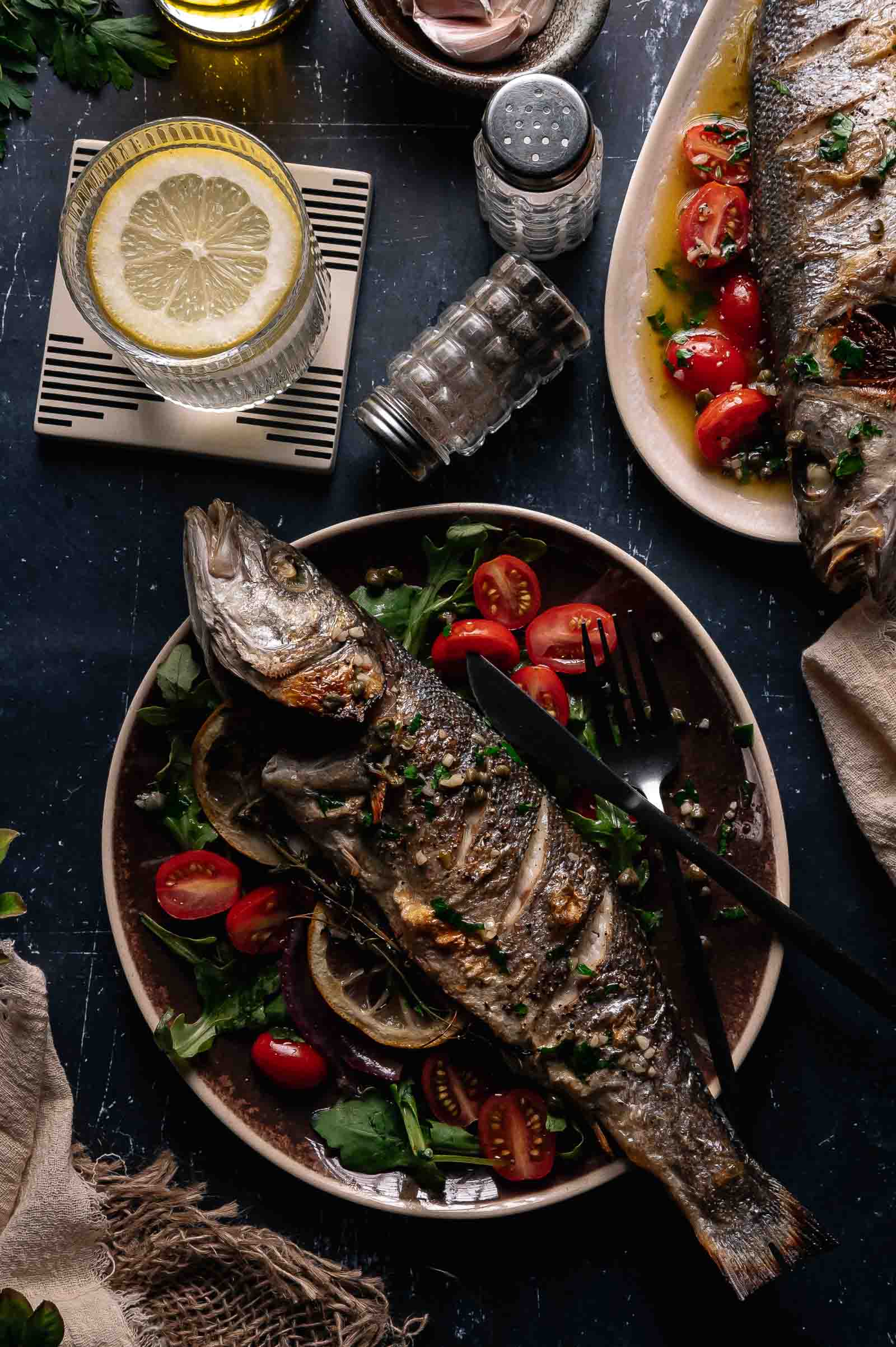 A whole roasted branzino served on a plate with mixed greens and cherry tomatoes, alongside a glass of lemon water and a salt shaker.
