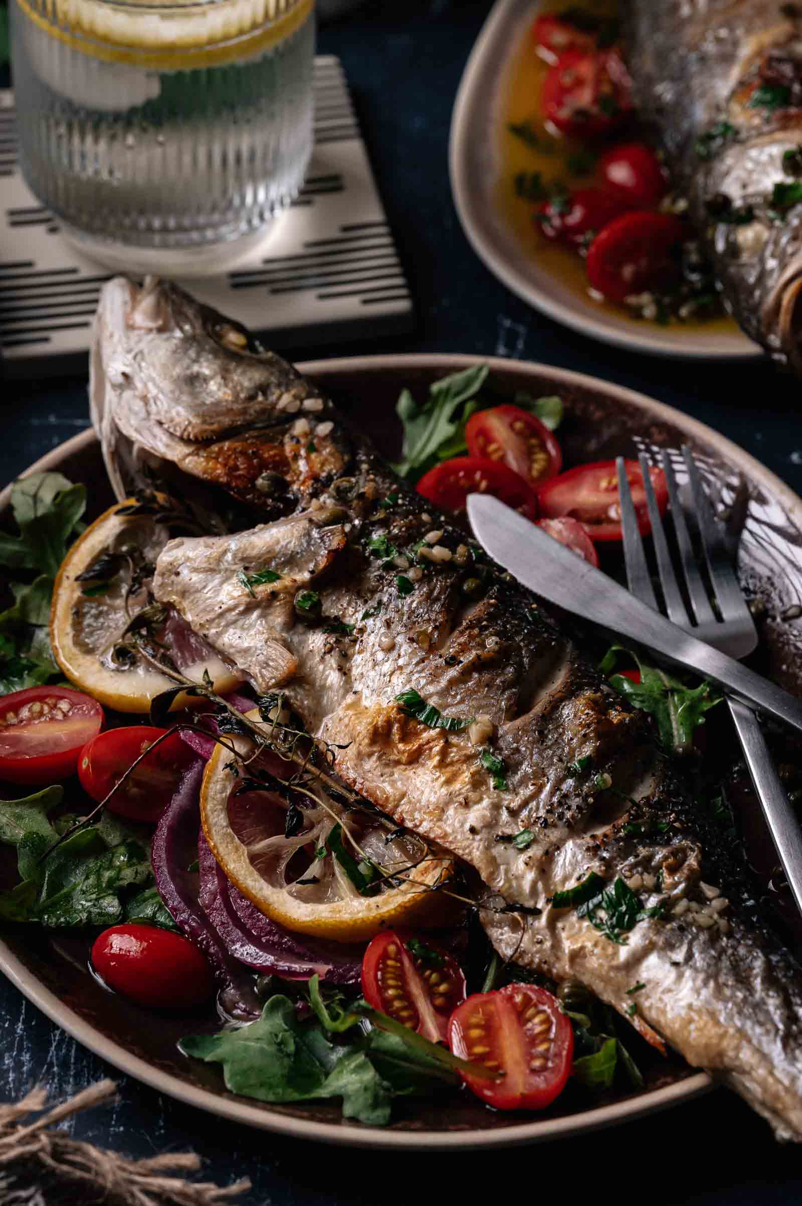 Whole roasted branzino served on a plate with lemon slices, cherry tomatoes, greens, and herbs; knife and fork placed beside the fish.