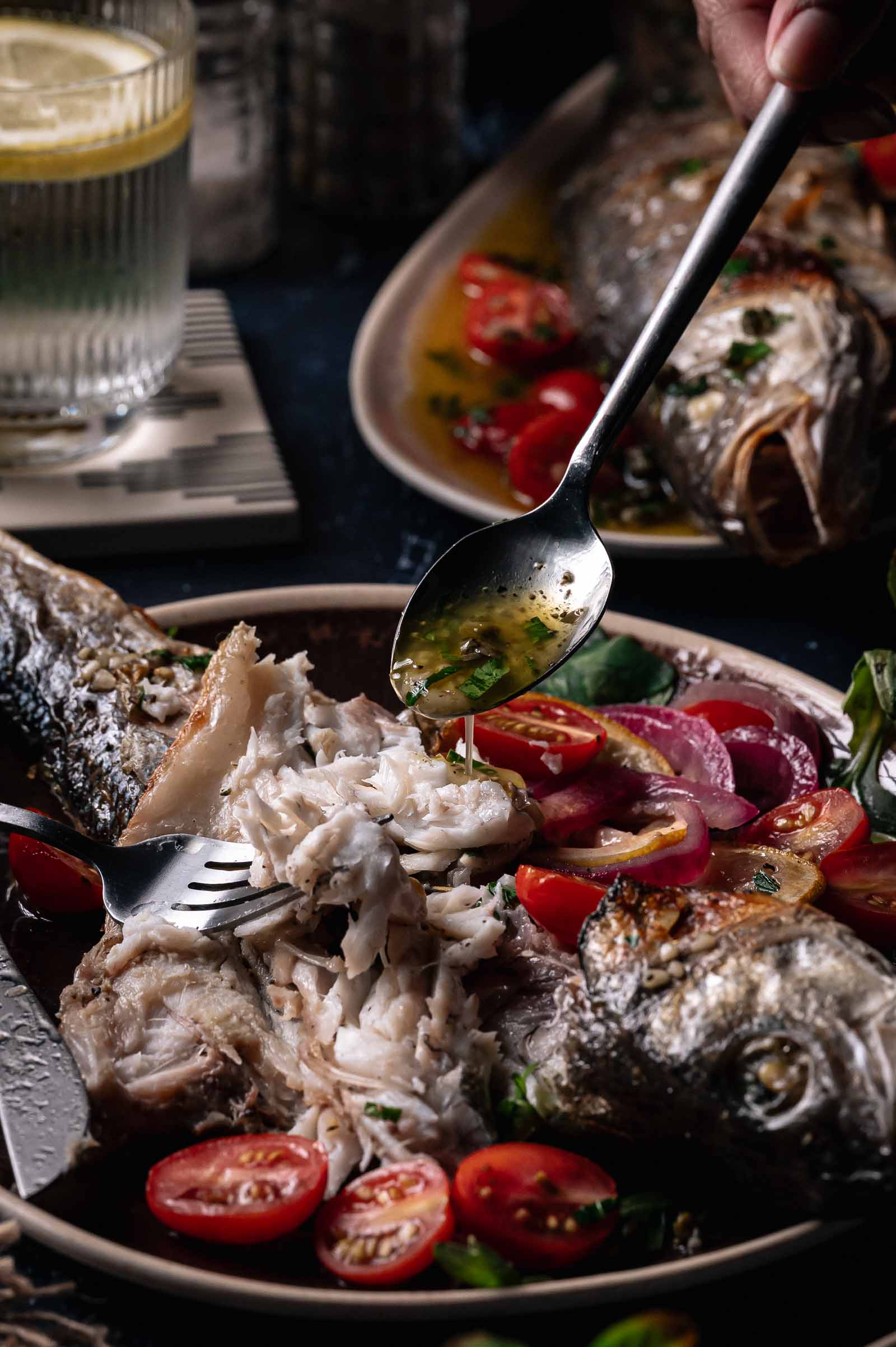 A cooked whole roasted branzino is being drizzled with sauce, with a spoon drizzling sauce over it. Sliced cherry tomatoes and greens are served on the side.