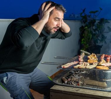 A man holds his head in frustration while grilling assorted meats on a barbecue, with smoke rising from the grill.