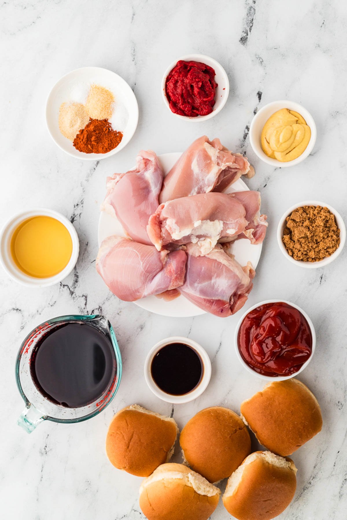 Raw chicken thighs on a plate surrounded by ingredients in bowls, including spices, ketchup, tomato paste, mustard, brown sugar, red wine, Worcestershire sauce, oil, and sandwich buns.