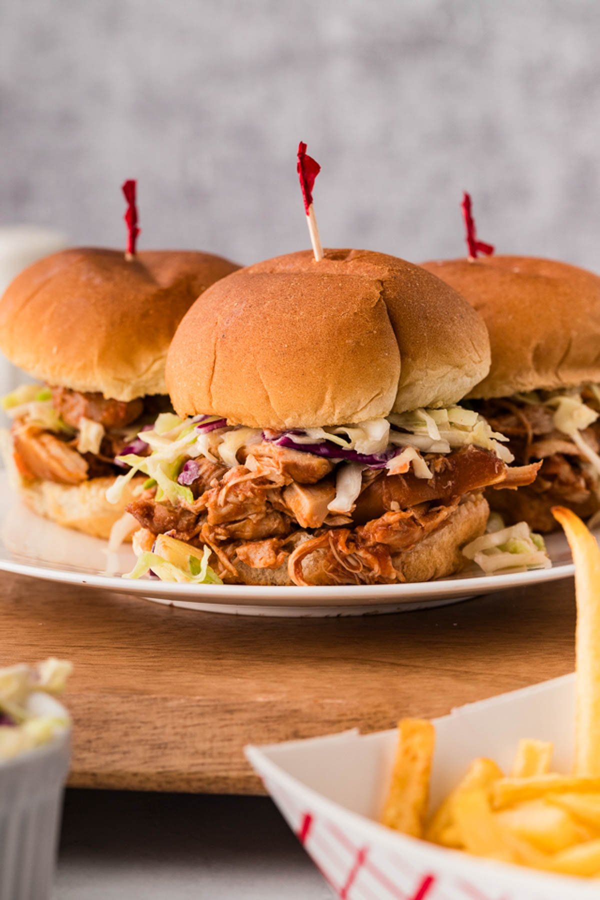 Three bbq chicken sliders topped with coleslaw on a white plate, with a side of French fries in the foreground.
