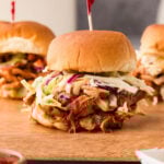 BBQ chicken slider topped with coleslaw on a wooden board, with two more sliders in the background.