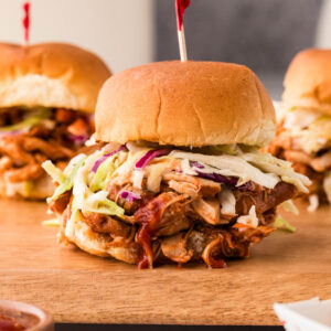 BBQ chicken slider topped with coleslaw on a wooden board, with two more sliders in the background.