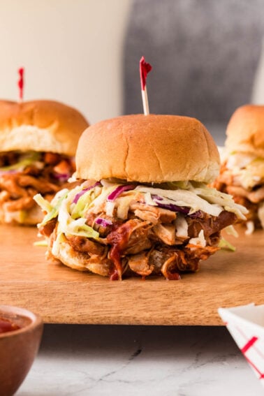 BBQ chicken slider topped with coleslaw on a wooden board, with two more sliders in the background.