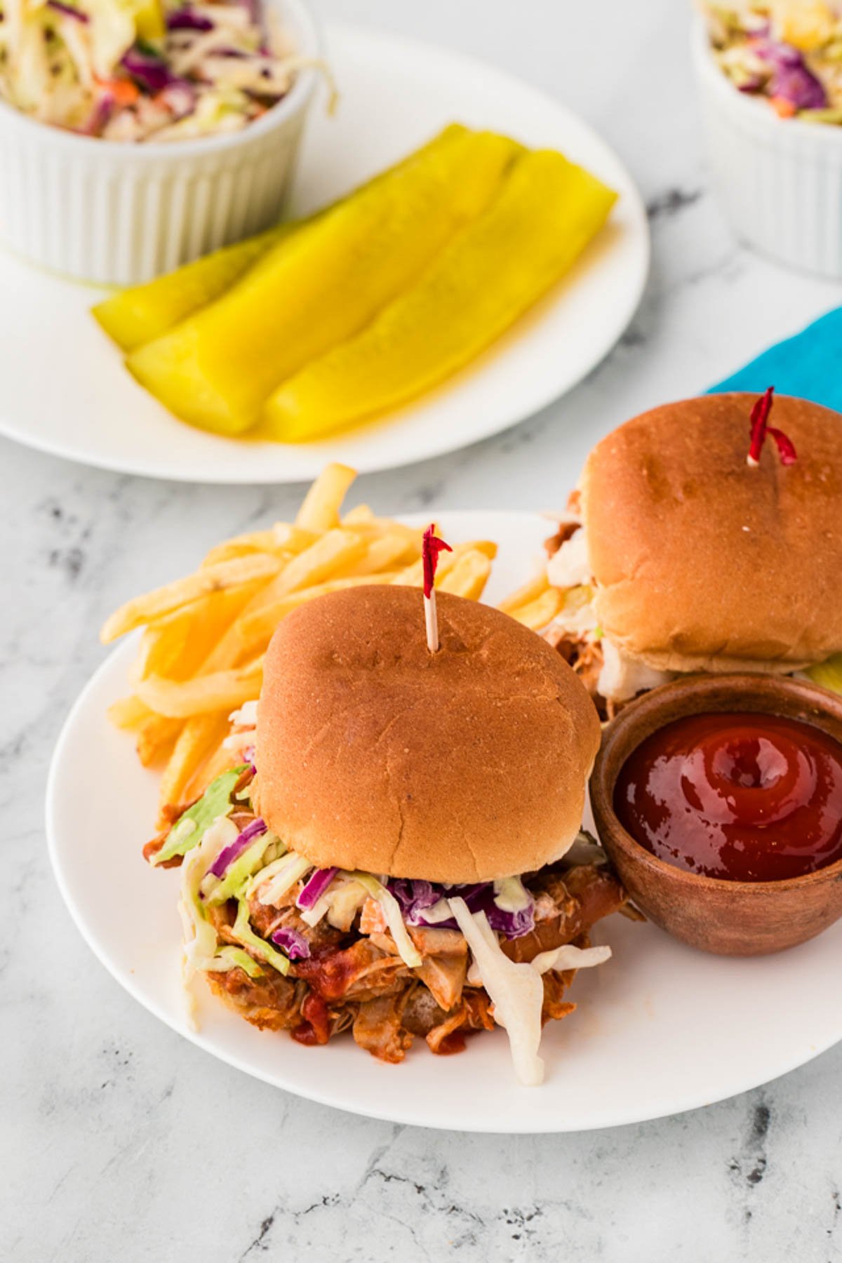 Two barbecue chicken sliders with coleslaw on a plate, served with French fries, ketchup, and a side of pickle spears in the background.