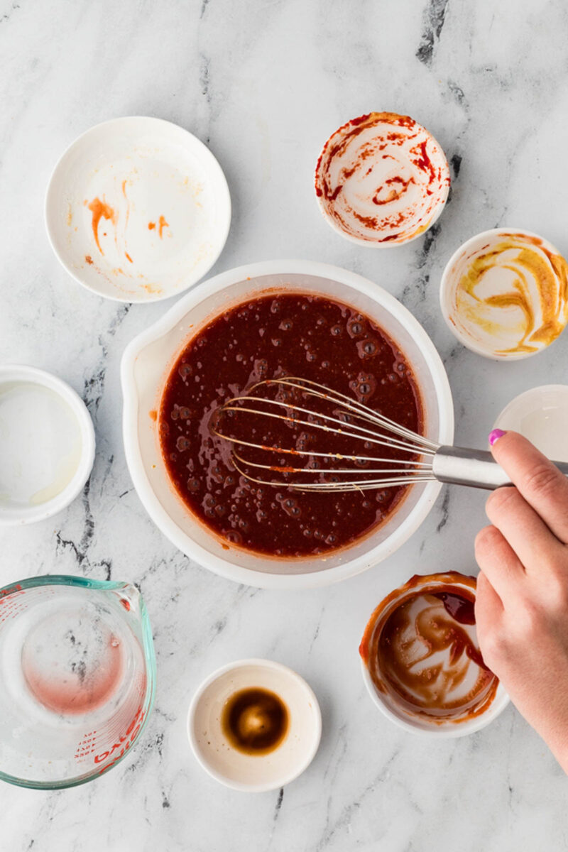 A hand whisks cherry bbq sauce in a bowl surrounded by empty and partially used ingredient bowls on a marble countertop.