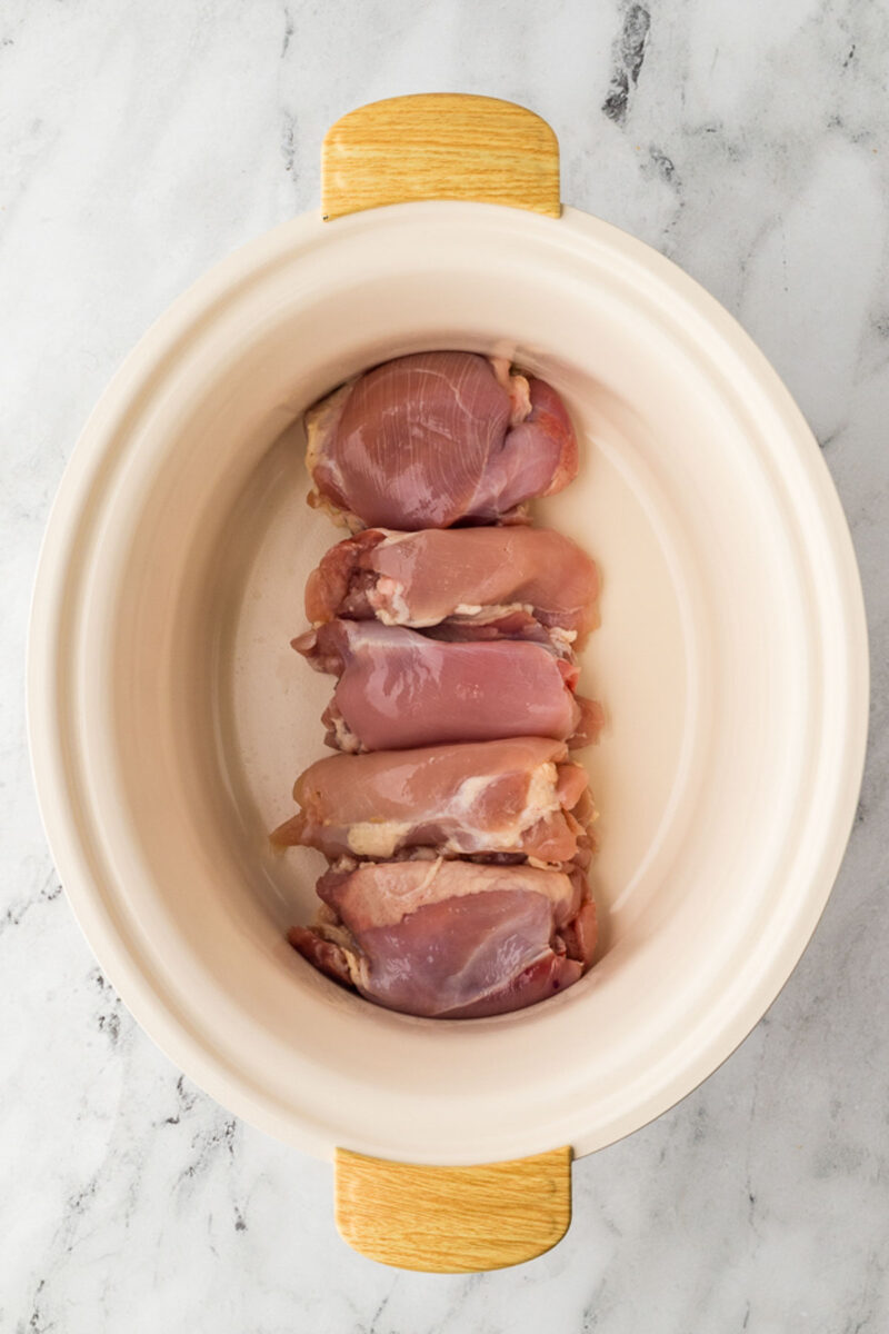 Raw boneless chicken thighs arranged in a single layer inside a cream-colored slow cooker on a marble surface.