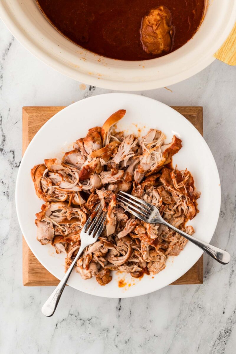 A white plate with shredded chicken and two forks, placed on a wooden board, with a slow cooker of sauce partially visible above.
