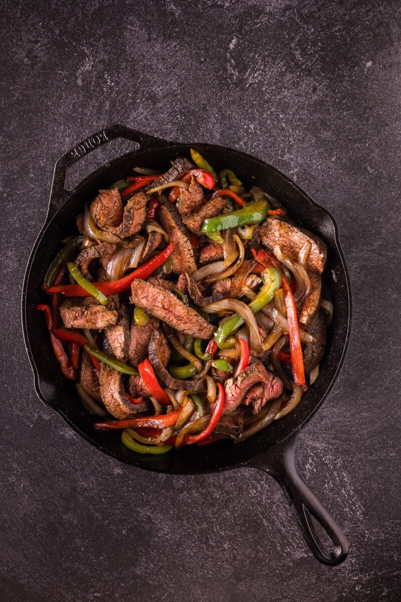 A cast iron skillet filled with cooked steak fajitas - beef strips, red and green bell peppers, and onions on a dark textured surface.