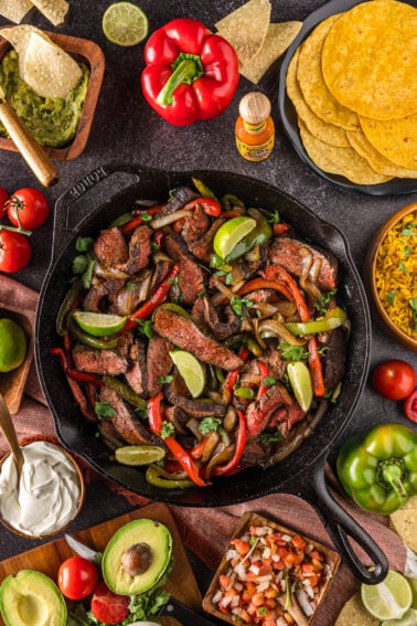 A skillet of beef steak fajitas with bell peppers and lime, surrounded by tortillas, guacamole, sour cream, pico de gallo, avocado, rice, chips, and fresh vegetables.