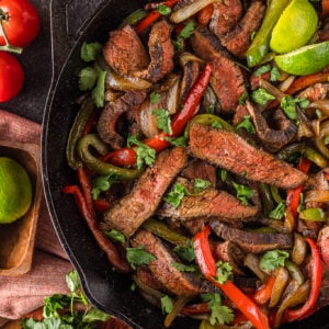 A cast iron skillet with sliced steak fajitas, red and green bell peppers, onions, and fresh cilantro, surrounded by tomatoes, limes, and avocado.