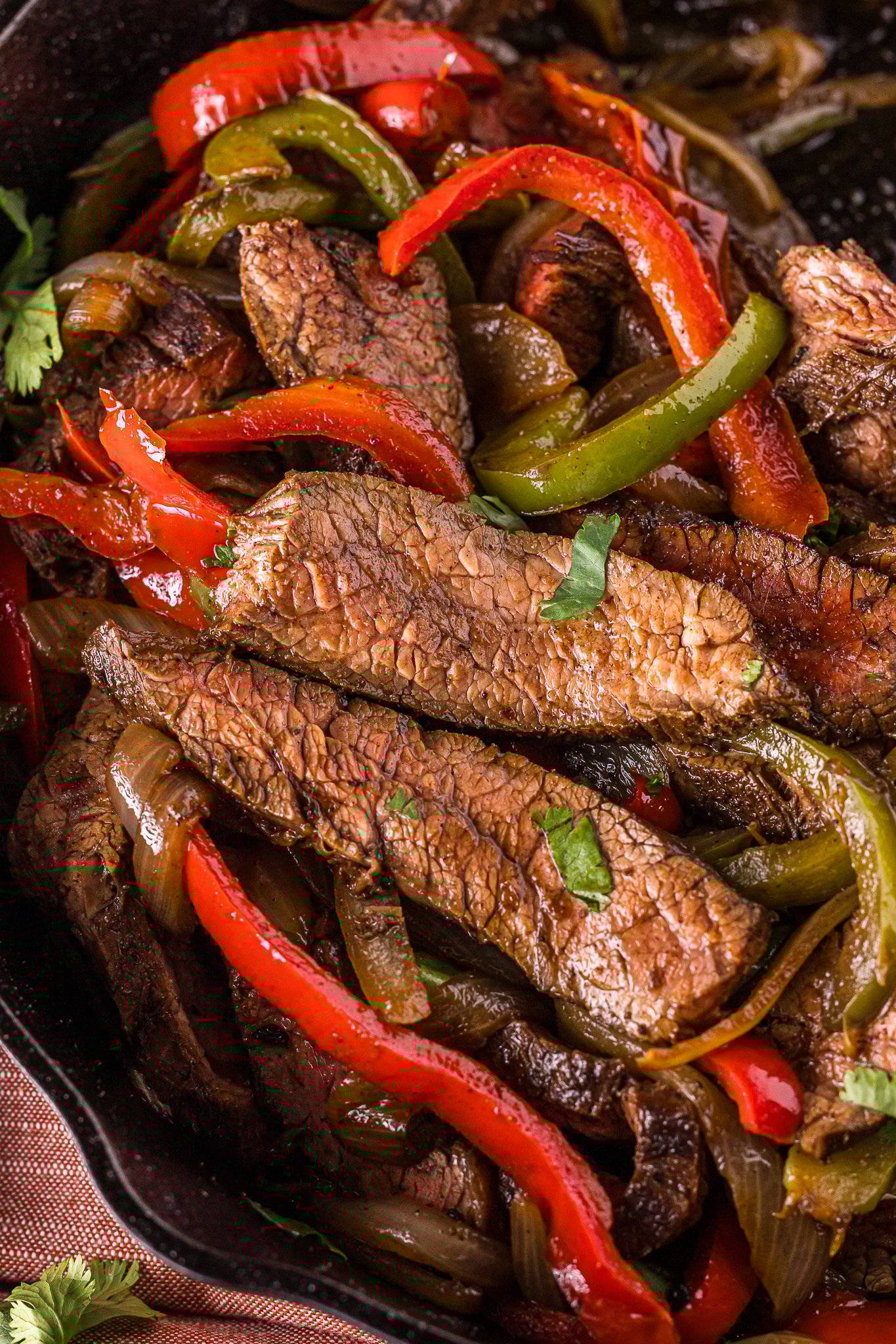 Close up of beef fajitas; Sliced cooked steak with red and green bell peppers and onions, garnished with cilantro, served in a skillet.