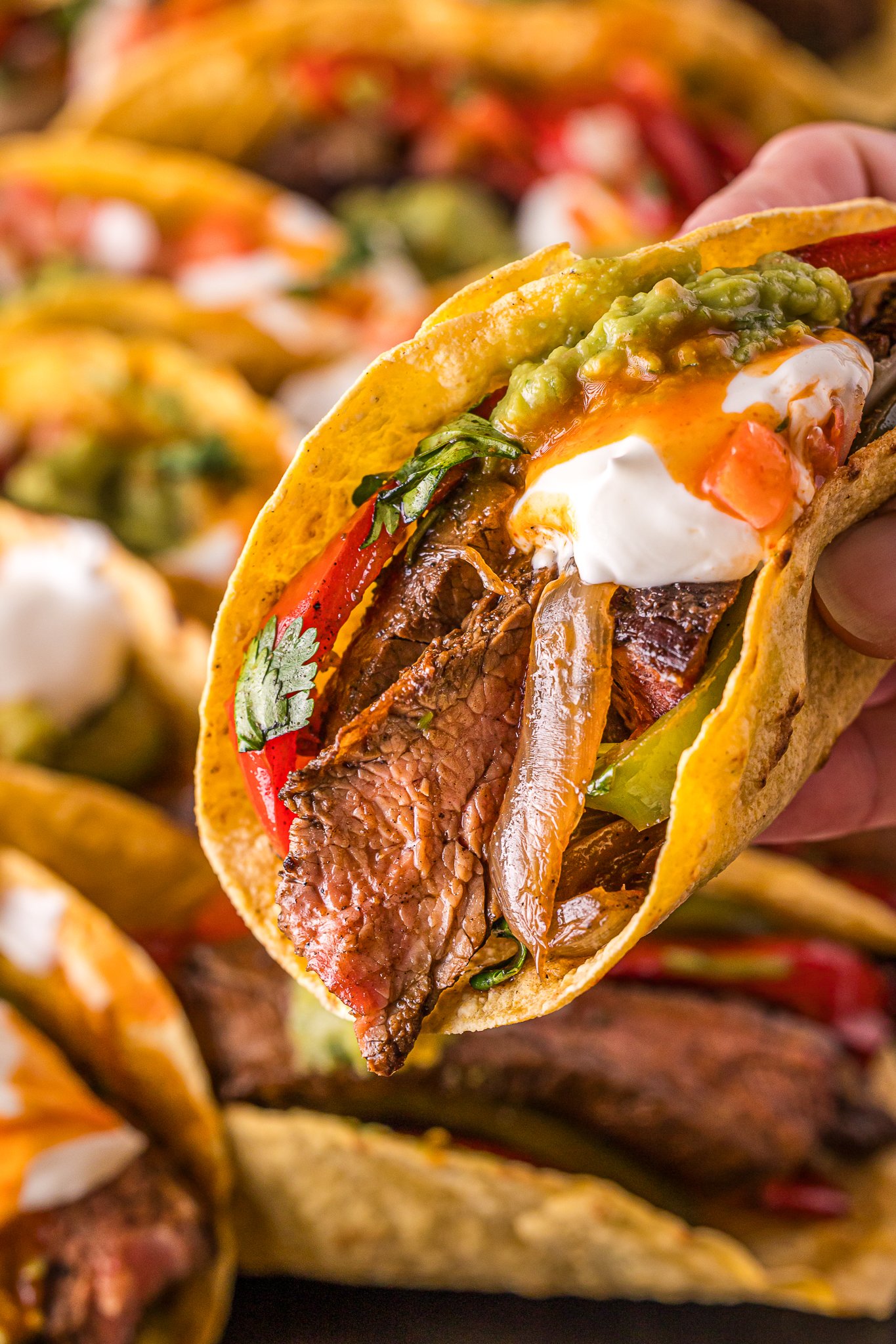 A hand holds a steak fajita with grilled steak, sautéed peppers and onions, guacamole, sour cream, salsa, and cilantro in a yellow corn tortilla.