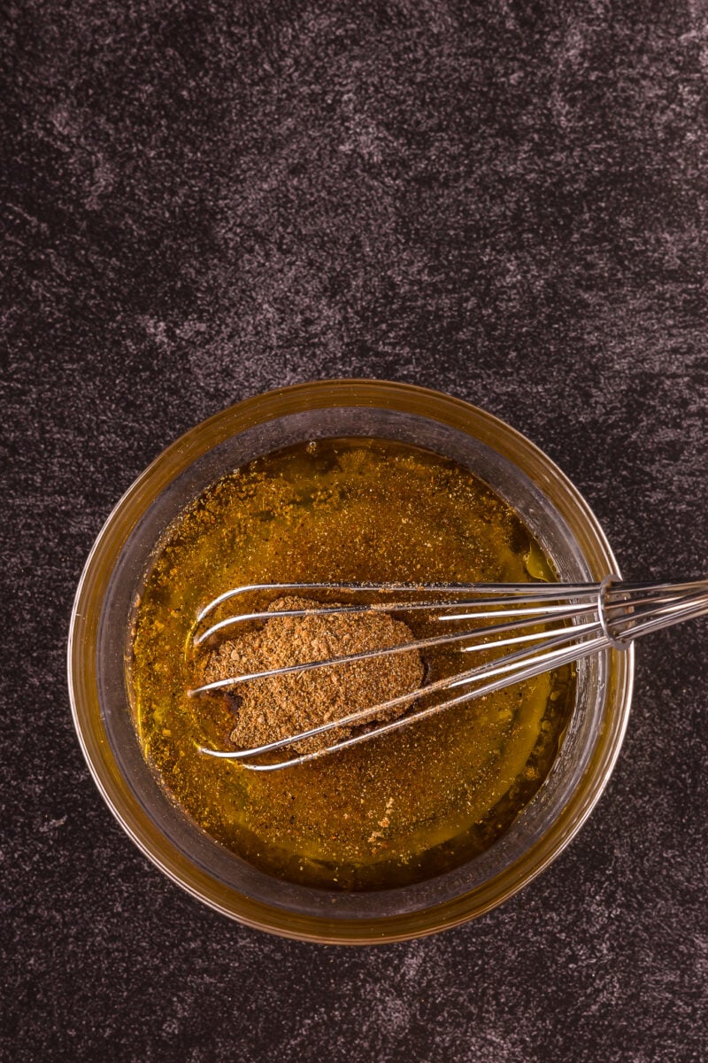A glass bowl with oil and spices being mixed with a metal whisk on a dark countertop.