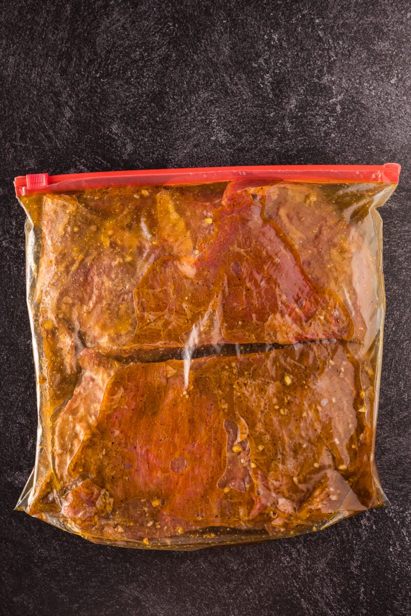 A sealed plastic bag containing pieces of flank steak marinating in a seasoned liquid, placed on a dark surface.