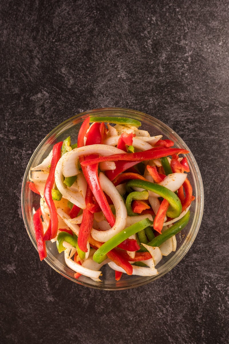 A glass bowl filled with sliced red and green bell peppers and onions, placed on a dark textured surface.