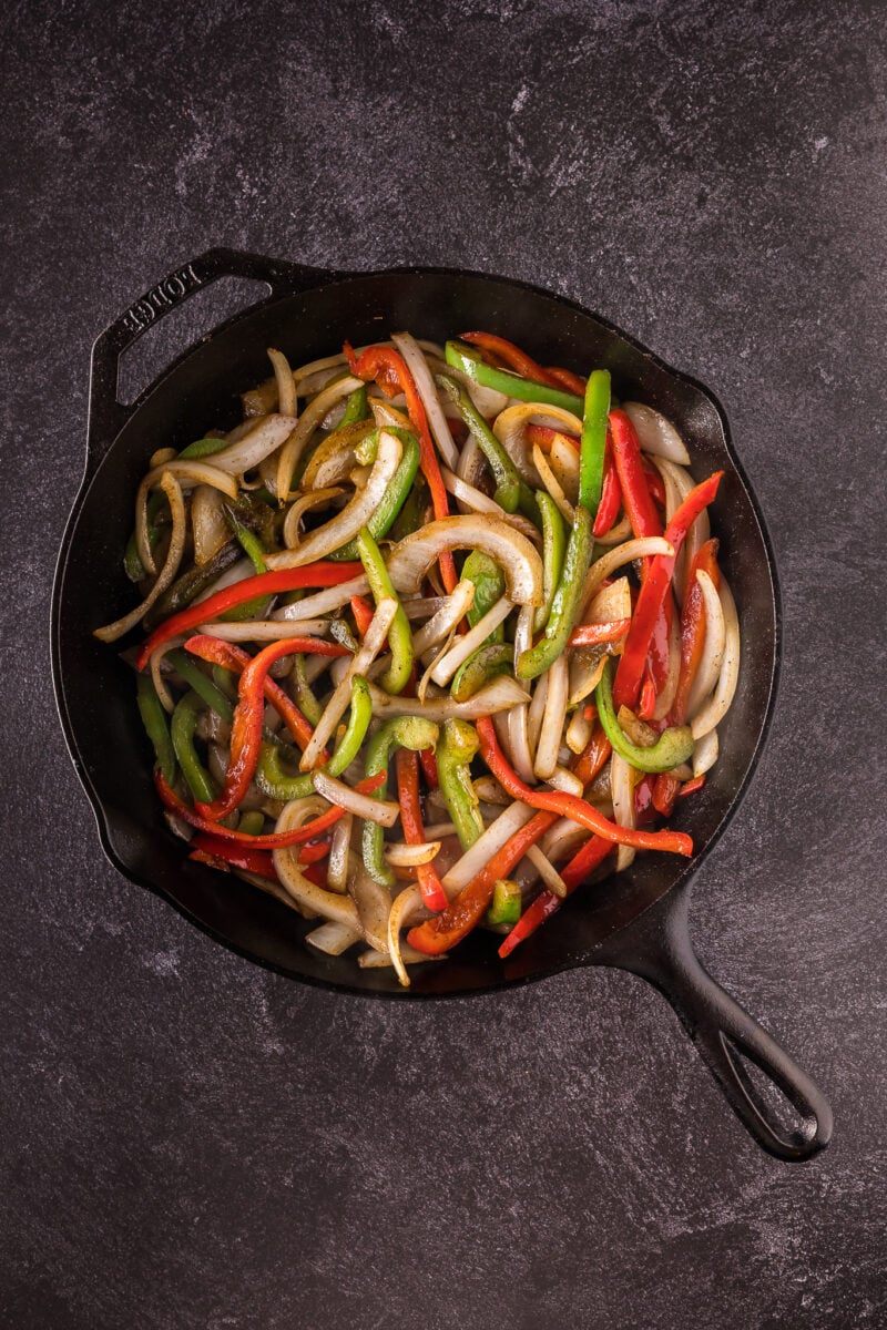 Sliced red and green bell peppers and onions sautéing in a black cast iron skillet on a dark textured surface.