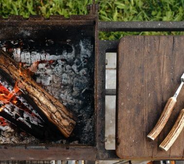A grill with burning logs on the left and a wooden board with a meat fork and carving knife on the right, set outdoors on grass.