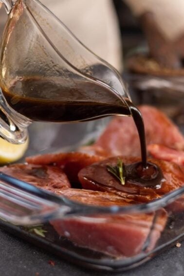 A person pours dark marinade over raw meat in a glass baking dish, surrounded by lemon and lime wedges, oil, salt, pepper, and herbs on a black countertop.