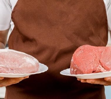 A person wearing a brown apron holds a plate with a piece of raw pork in one hand and a plate with a piece of raw beef in the other.