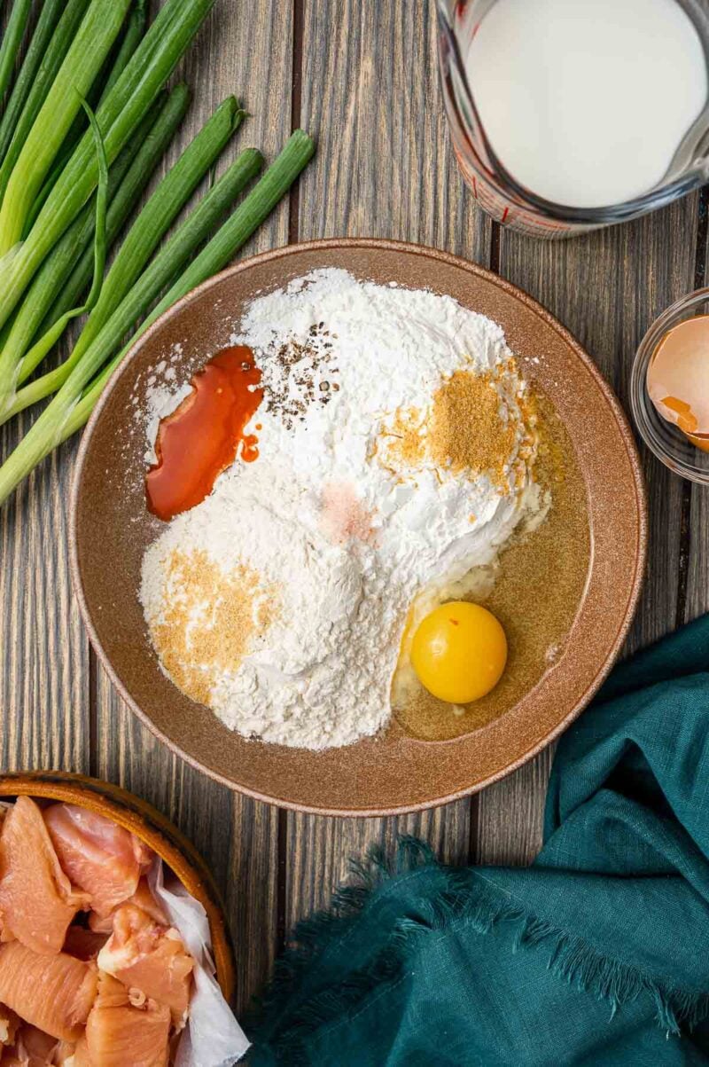 A bowl with flour, seasoning, an egg, and hot sauce sits on a wooden surface near chicken pieces, green onions, a cracked egg, and a glass of milk.