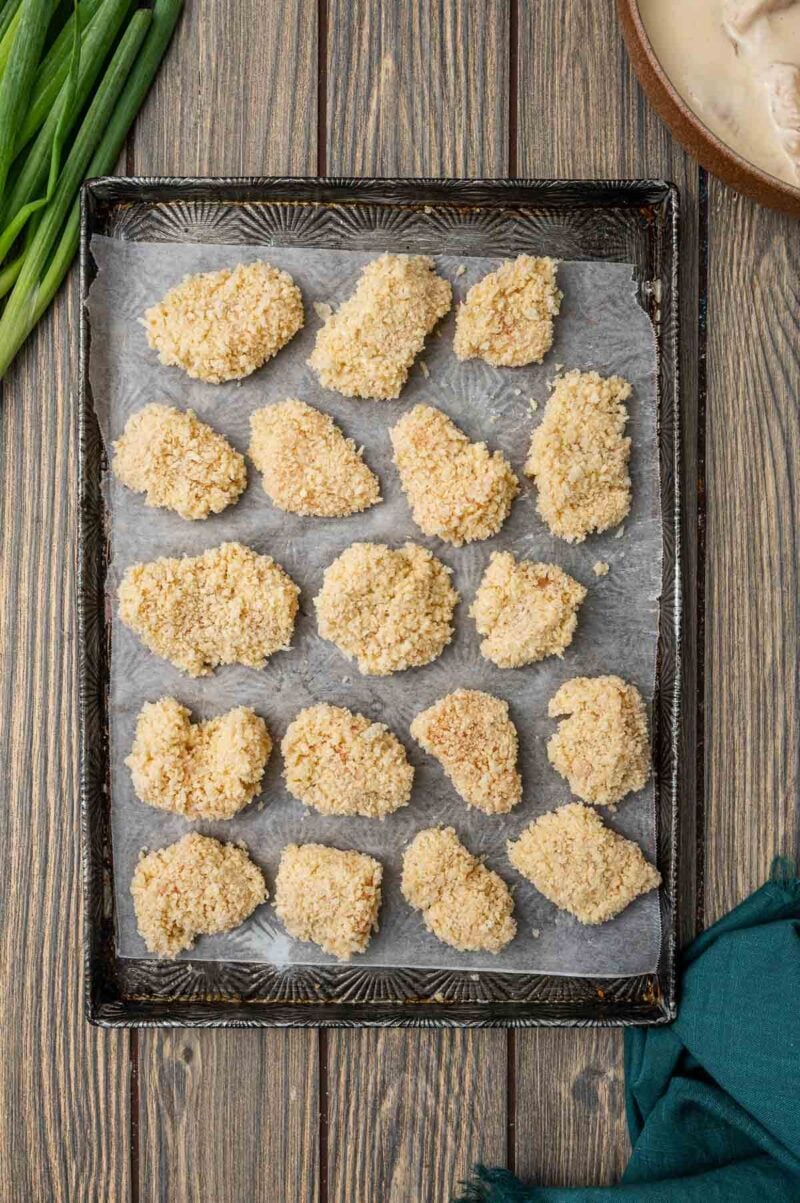 A baking tray lined with parchment paper holds sixteen evenly spaced, breaded chicken pieces ready to be cooked. Green onions and a bowl are nearby on a wooden surface.