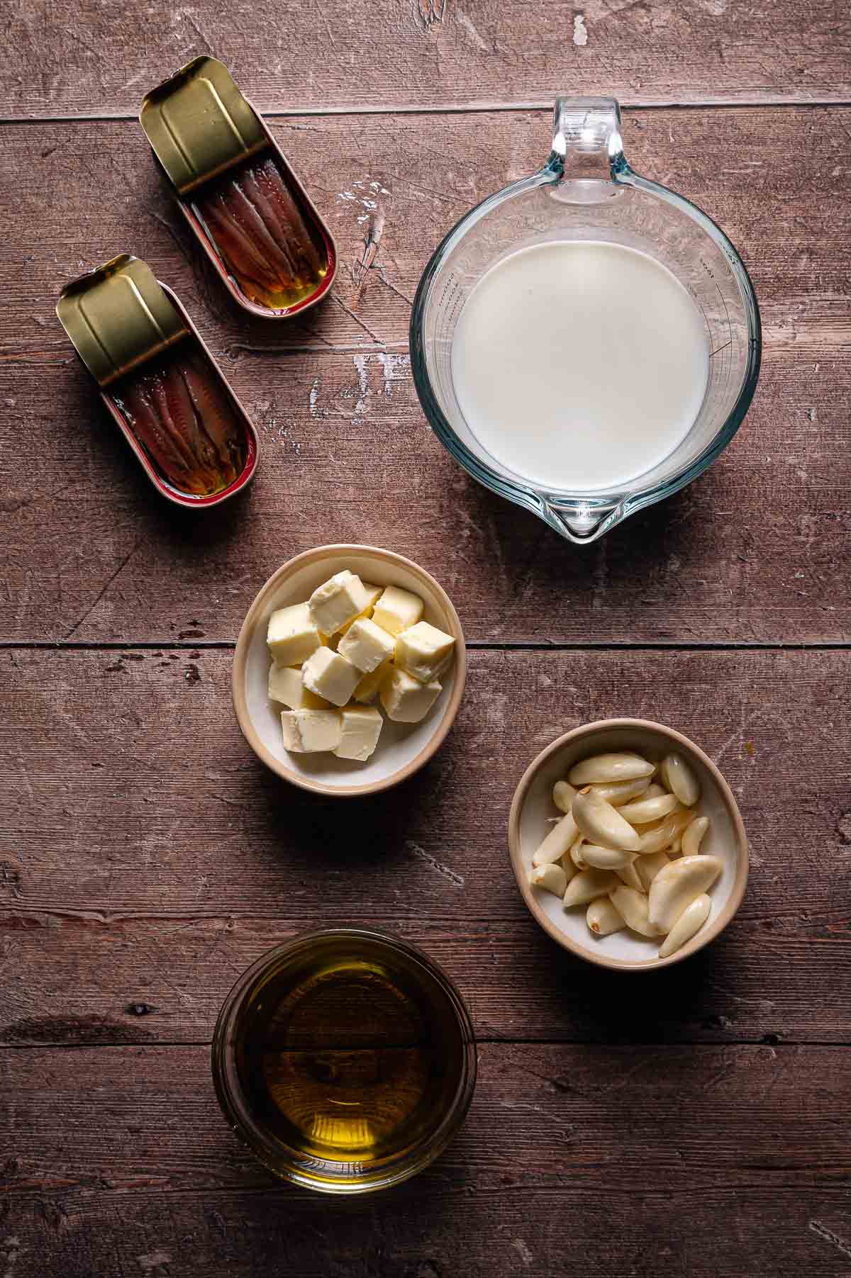 Overhead view of ingredients on a wooden surface: two tins of anchovies, a measuring cup of milk, a bowl of cubed butter, a bowl of garlic cloves, and a bowl of olive oil.
