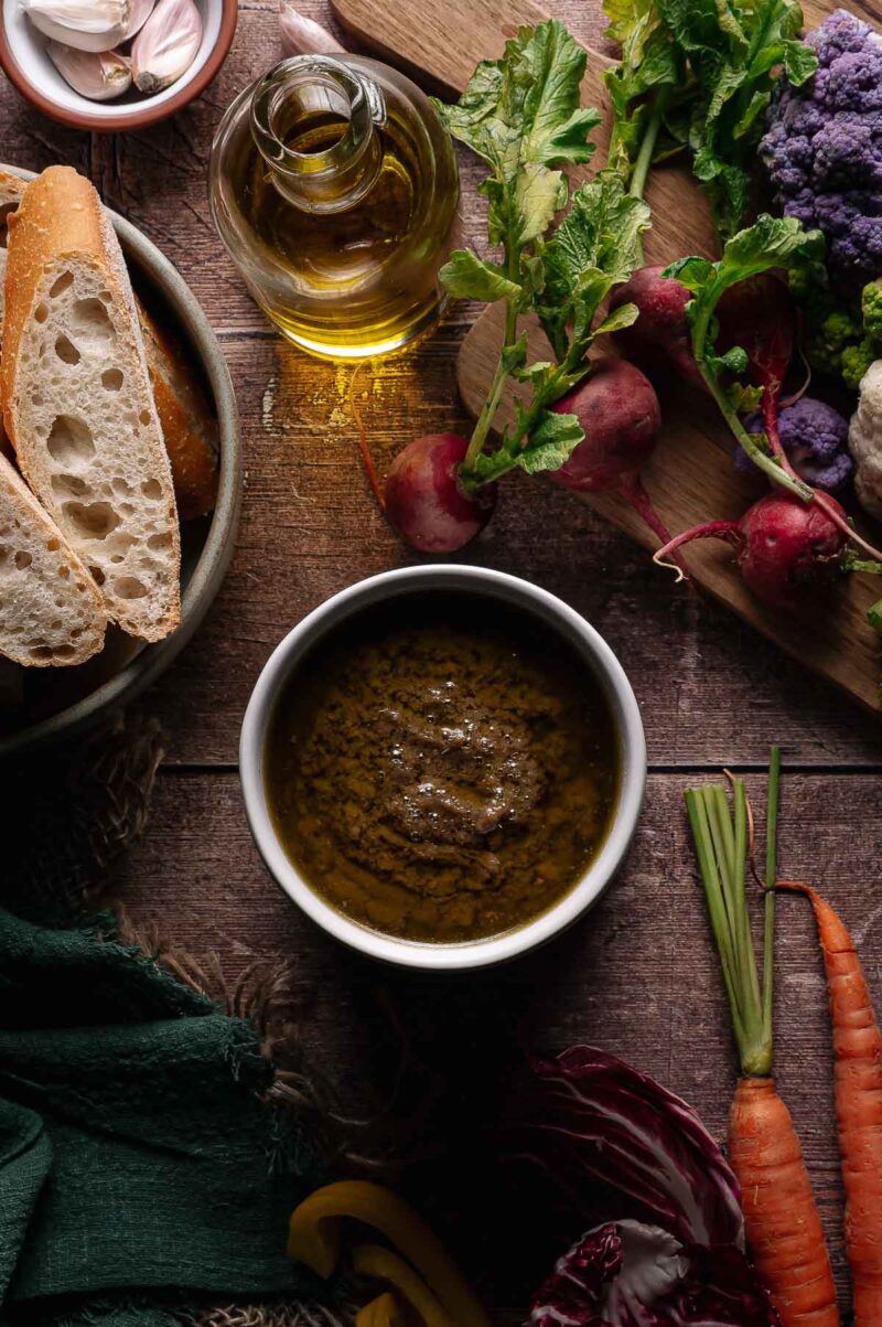 A bowl of Bagna cauda dip surrounded by sliced baguette, olive oil, radishes, carrots, cauliflower, and leafy greens on a wooden surface.