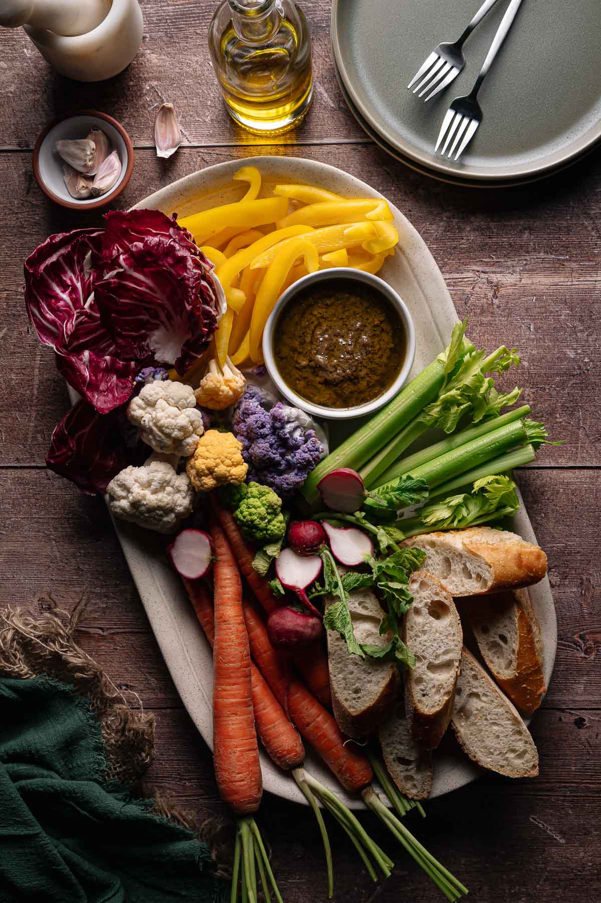 A platter with sliced baguette, carrots, radishes, celery, cauliflower, yellow bell pepper, radicchio, and a bowl of Bagna cauda dip, set on a wooden table with plates, forks, and olive oil.