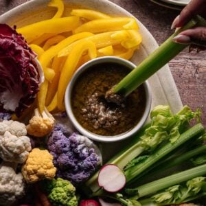 A platter with assorted raw vegetables, including yellow bell peppers, cauliflower, carrots, celery, and radishes, surrounds a bowl of Bagna cauda dip; a hand dips celery into the bowl.